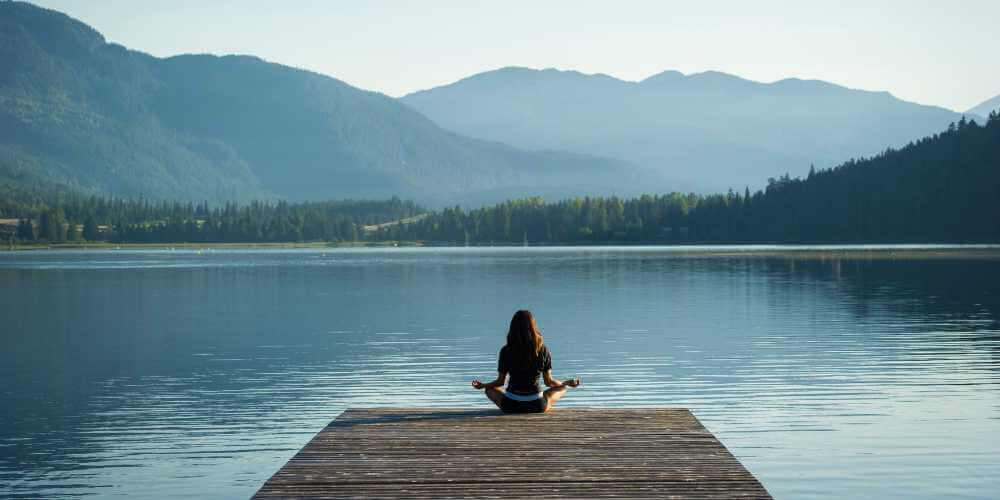 Person im Lotus-Sitz auf Steg am See – Symbol für Selbstfürsorge, innere Ruhe und achtsame Auszeit
